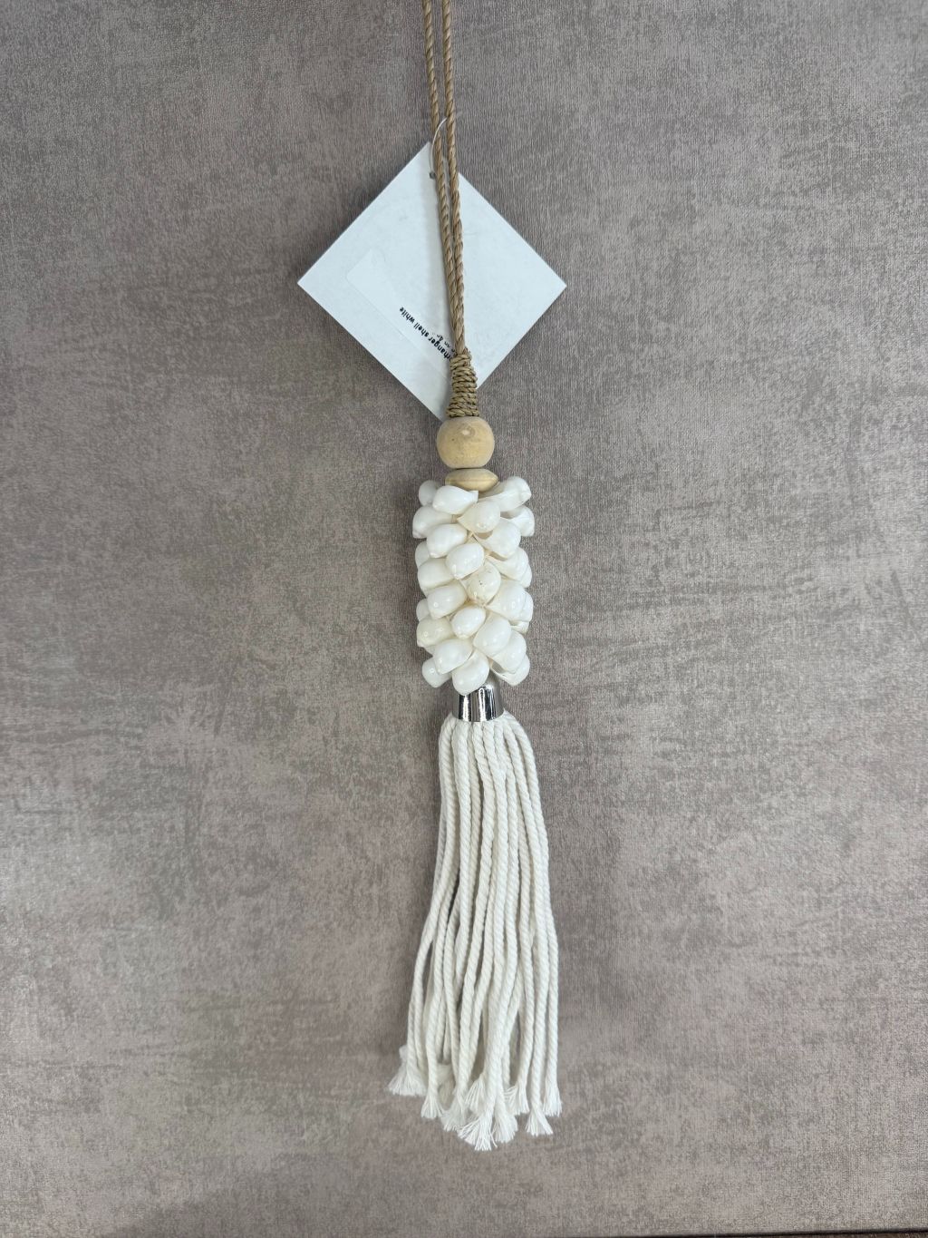 Witte hanger van schelpen en katoen met houten kraal en touw, hangend tegen een neutrale achtergrond.