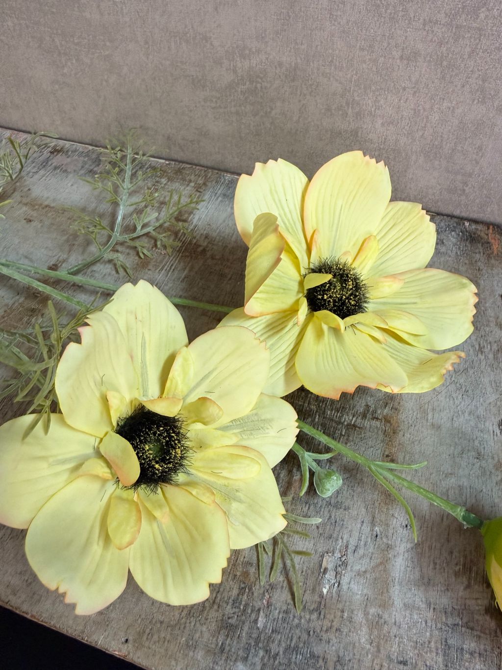 Gele kunstbloem anemoon met donkere harten en groene takjes tegen een neutrale achtergrond.