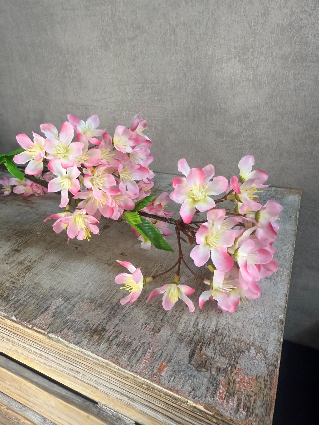 Lange kunsttak met vijf trosjes roze bloesem en groene blaadjes, tegen een grijze achtergrond.