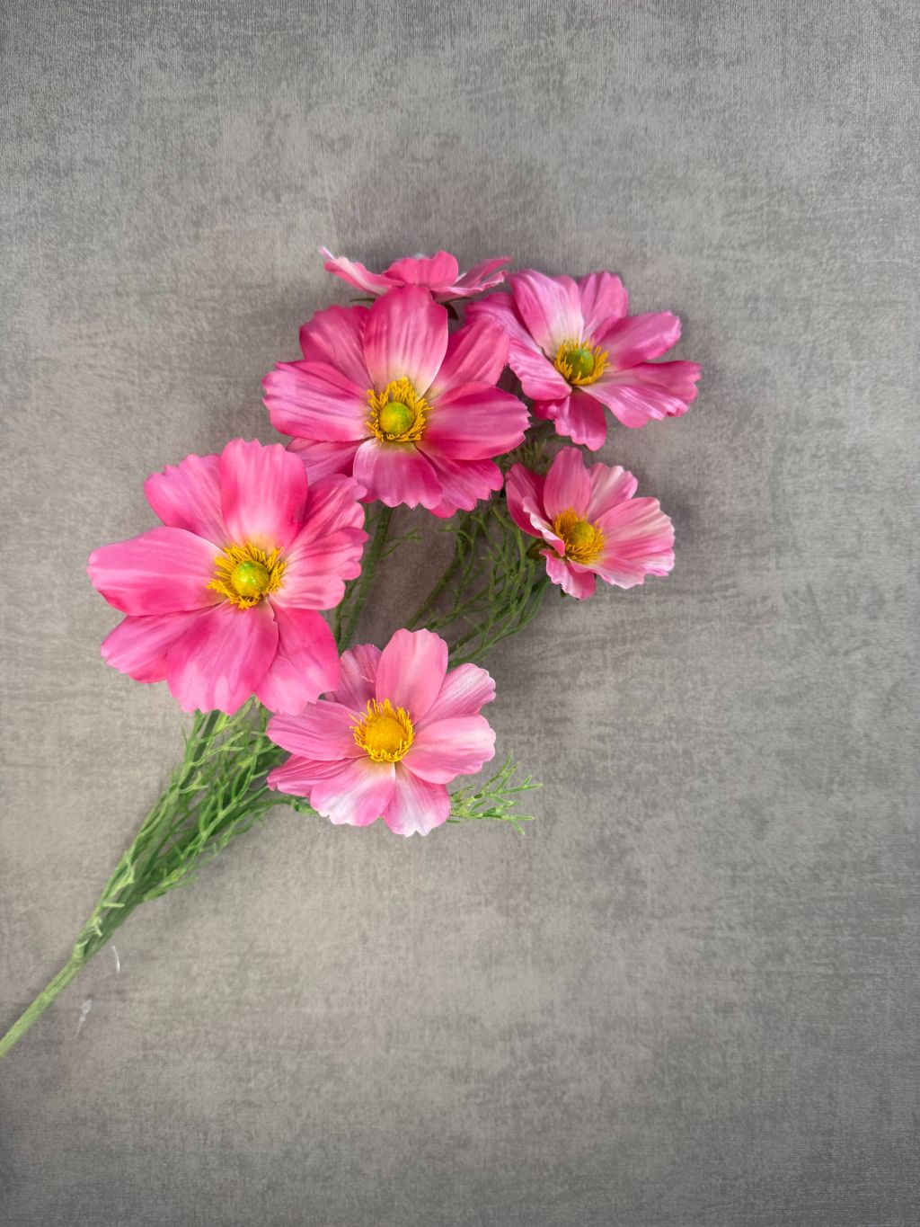 Kunstbloem Cosmos met zes roze bloemen met gele hartjes, op een groene steel met fijne blaadjes, liggend op een grijze ondergrond.