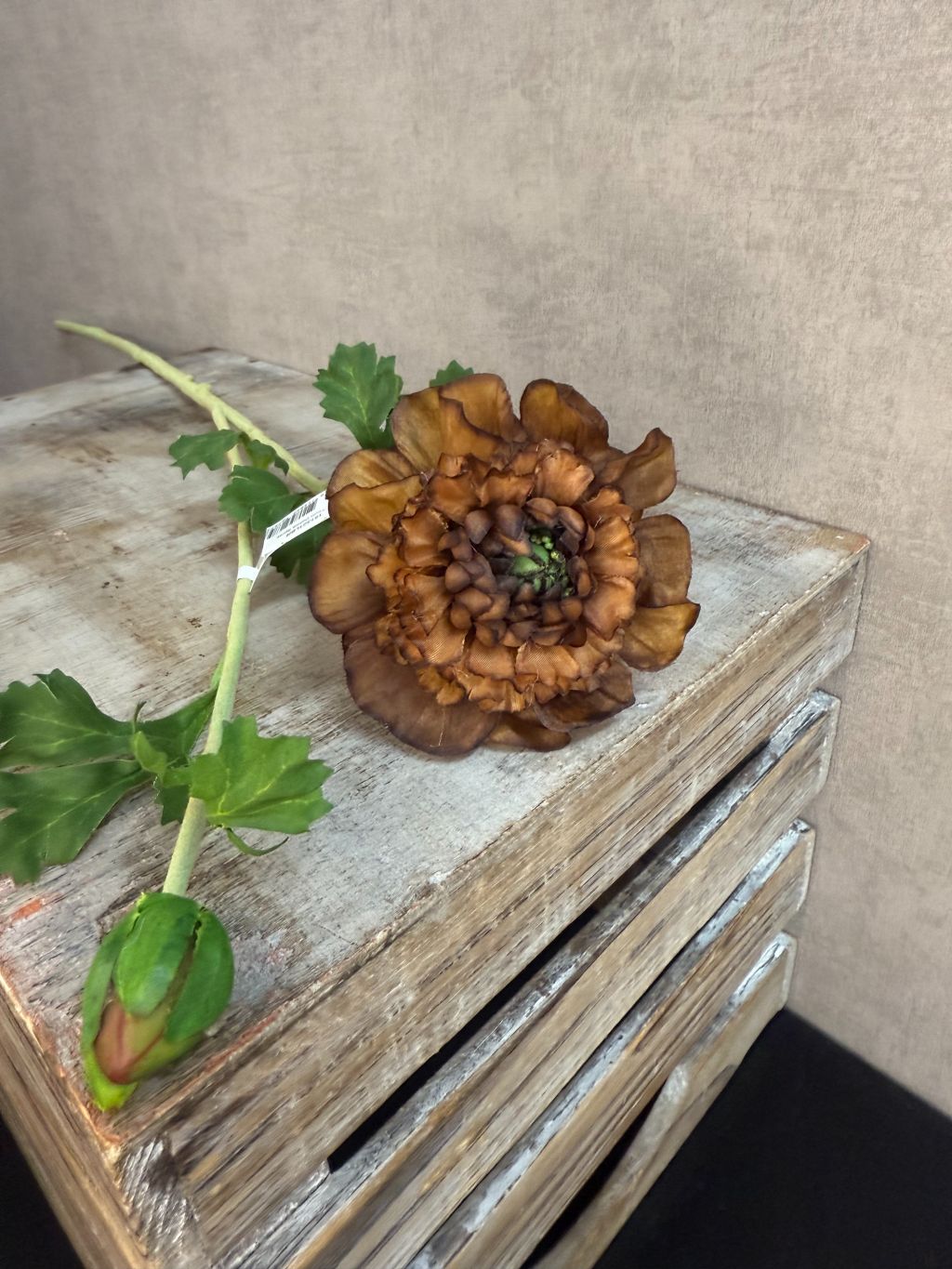 Bruine kunstbloem dahlia met lange steel en groene bladeren op een houten kist.