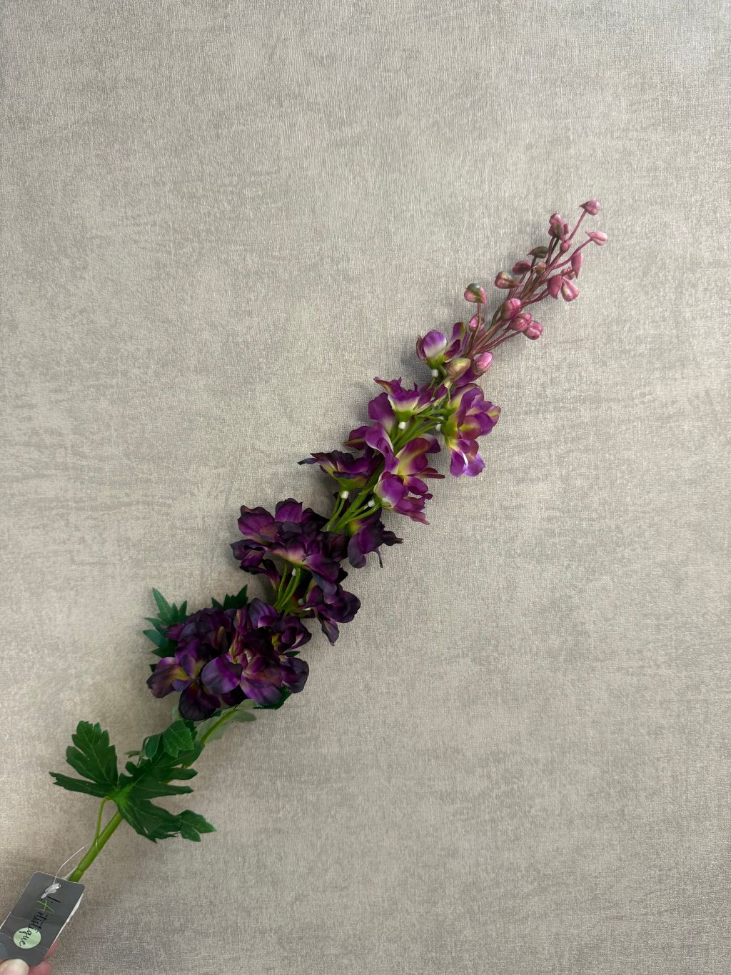Kunstbloem Delphinium Paars melange met paarse bloemen en groene steel, liggend op een grijze ondergrond.