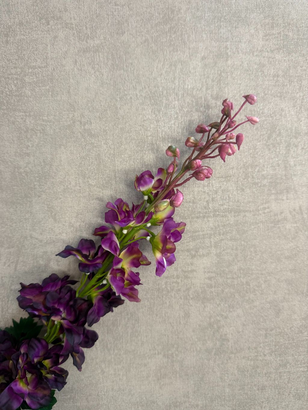 Kunstbloem Delphinium Paars melange met paarse bloemen en groene steel, liggend op een grijze ondergrond.
