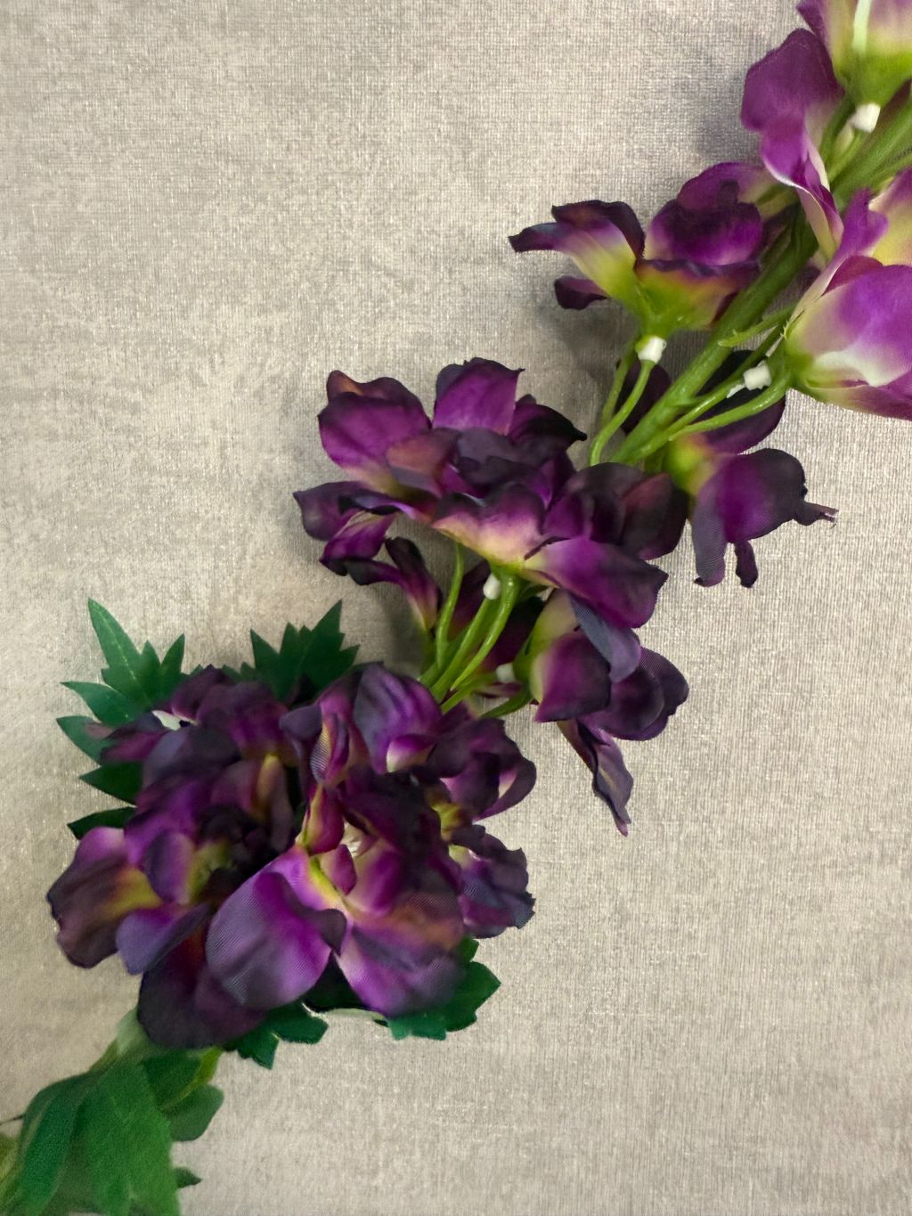 Kunstbloem Delphinium Paars melange met paarse bloemen en groene steel, liggend op een grijze ondergrond.