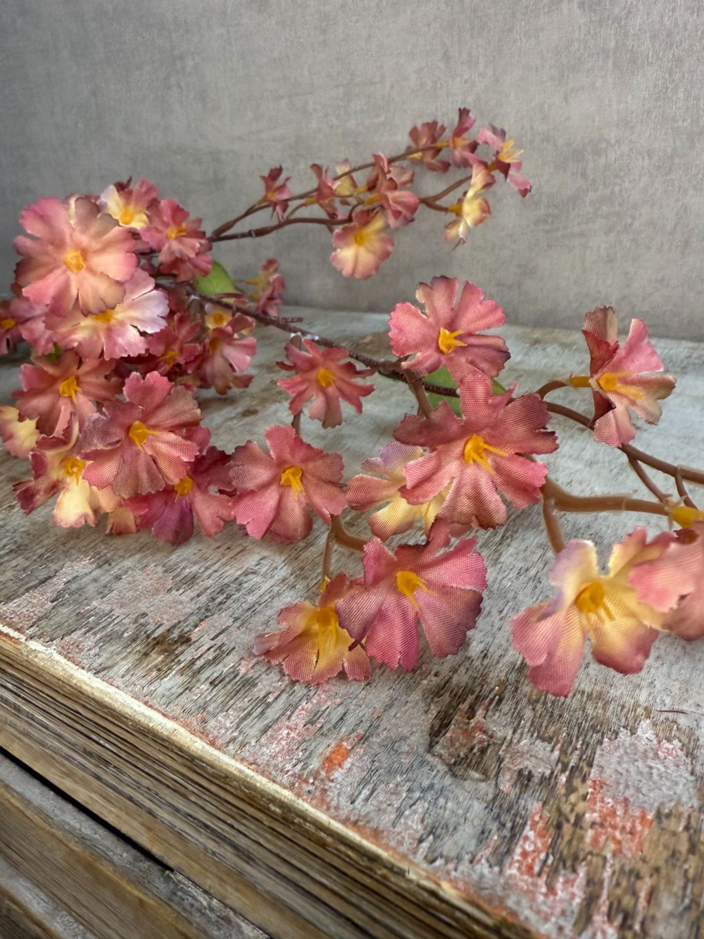 Kunsttak jasmijn in mauve tint met fijne bloemetjes en groene blaadjes op een donkerbruine steel, liggend op een grijze achtergrond.