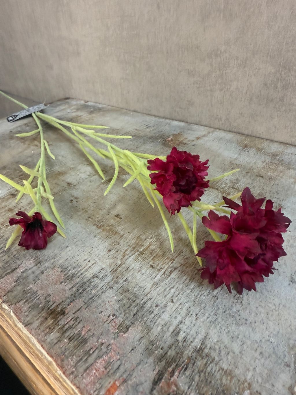 Kunstbloem korenbloem in bordeauxrood met drie bloemen aan groene steel, liggend op een grijze achtergrond.