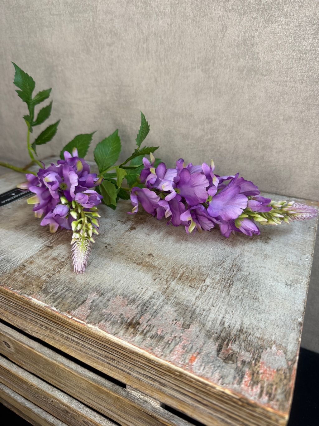 Kunstbloem lupine in het paars met groene bladeren en dubbele bloemtros, gefotografeerd tegen een neutrale grijze achtergrond.