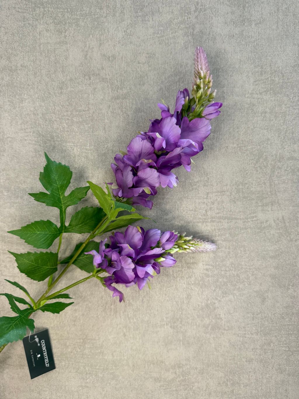 Kunstbloem lupine in het paars met groene bladeren en dubbele bloemtros, gefotografeerd tegen een neutrale grijze achtergrond.