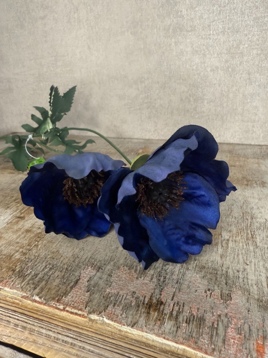 Donkerblauwe kunstklaproos met twee bloemen en groene bladeren tegen een houten ondergrond