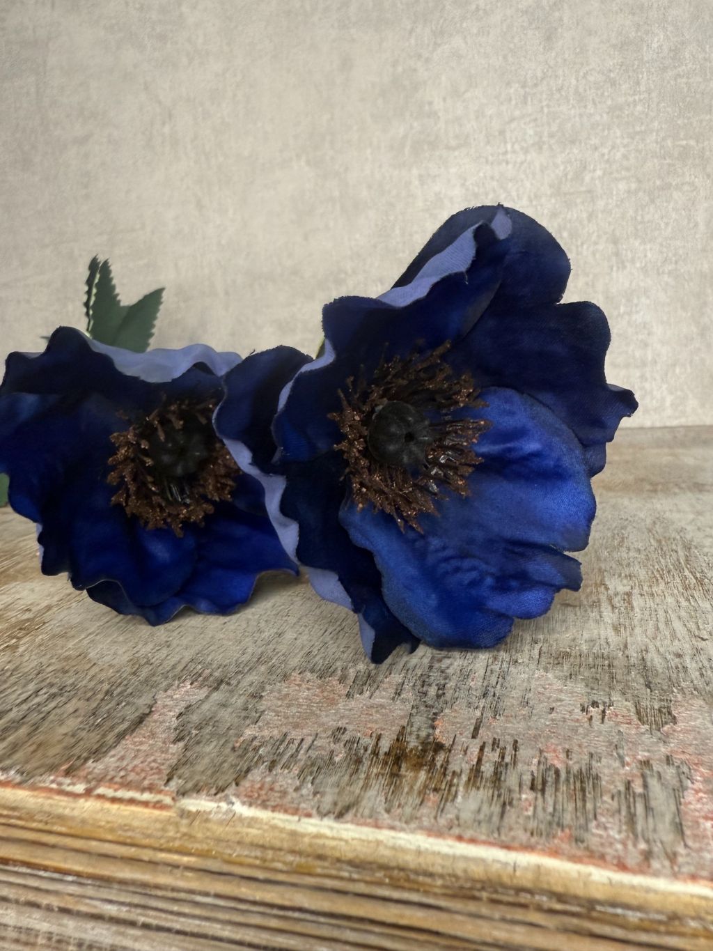 Donkerblauwe kunstklaproos met twee bloemen en groene bladeren tegen een houten ondergrond