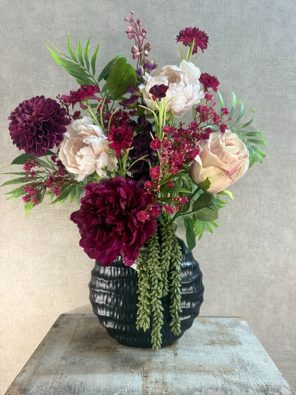 Kunstboeket Beau in zwarte ribbelvaas met bordeaux en roze bloemen, hangplant en groene accenten, front facing geschikt.