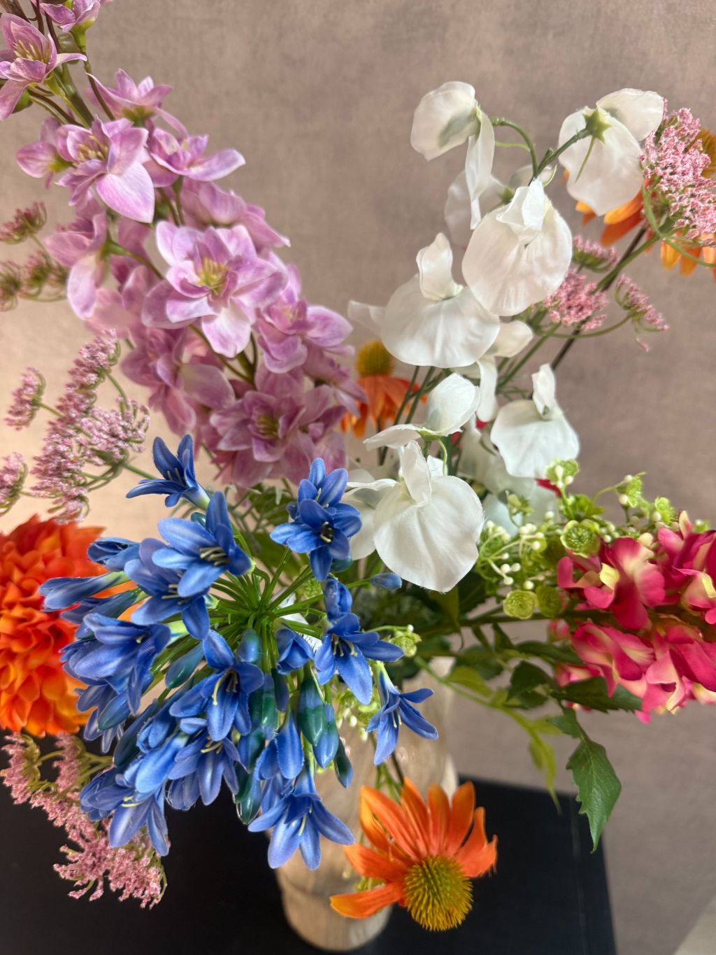 Kunstboeket met gemengde bloemen in diverse kleuren, geplaatst in een golvende beige vaas op zwarte ondergrond.