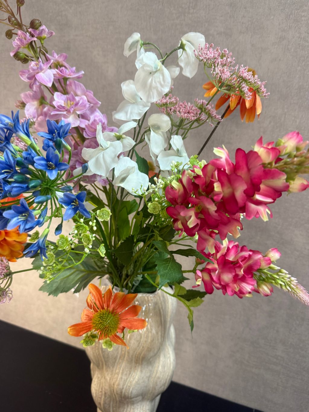 Kunstboeket met gemengde bloemen in diverse kleuren, geplaatst in een golvende beige vaas op zwarte ondergrond.