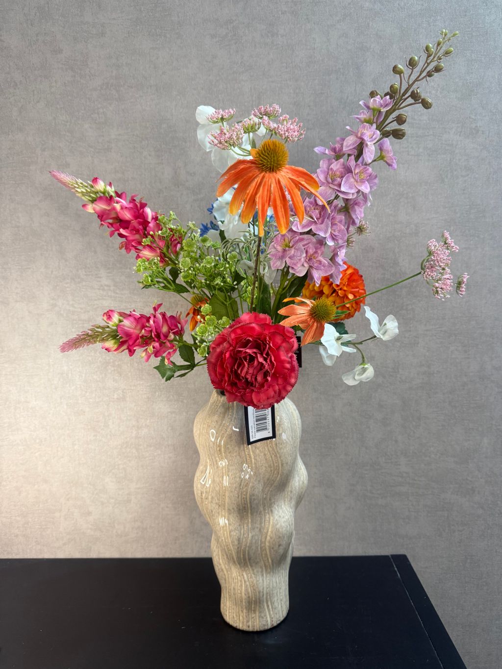 Kunstboeket met gemengde bloemen in diverse kleuren, geplaatst in een golvende beige vaas op zwarte ondergrond.