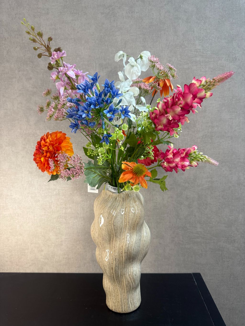 Kunstboeket met gemengde bloemen in diverse kleuren, geplaatst in een golvende beige vaas op zwarte ondergrond.