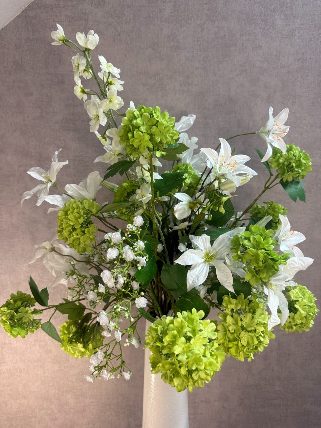 Kunstboeket met witte bloemen en groene sneeuwballen in een hoge crème vaas op zwarte ondergrond.