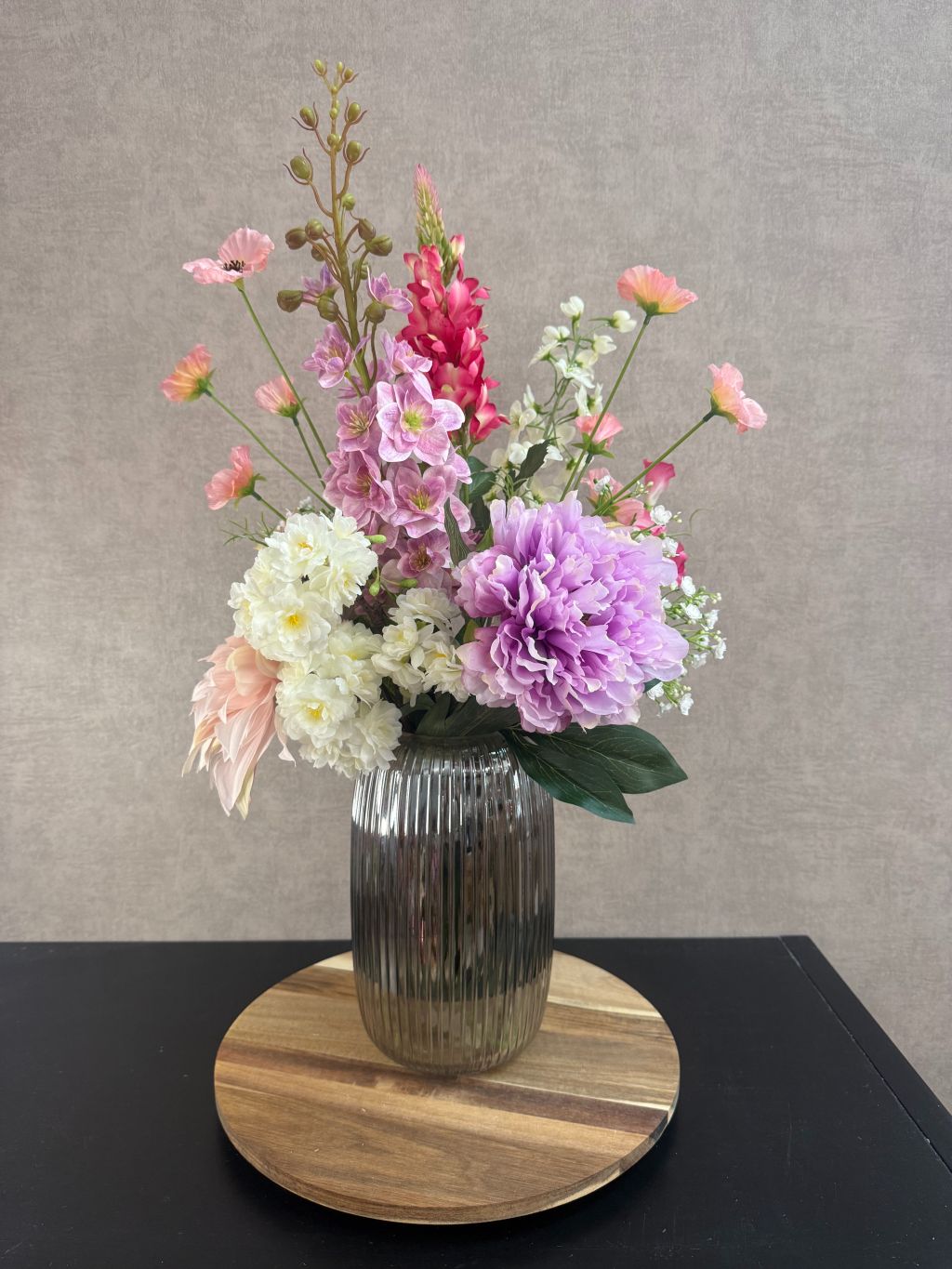 Kunstboeket Lovey met paarse, roze en witte bloemen in geribbelde glazen vaas op ronde houten onderzetter.