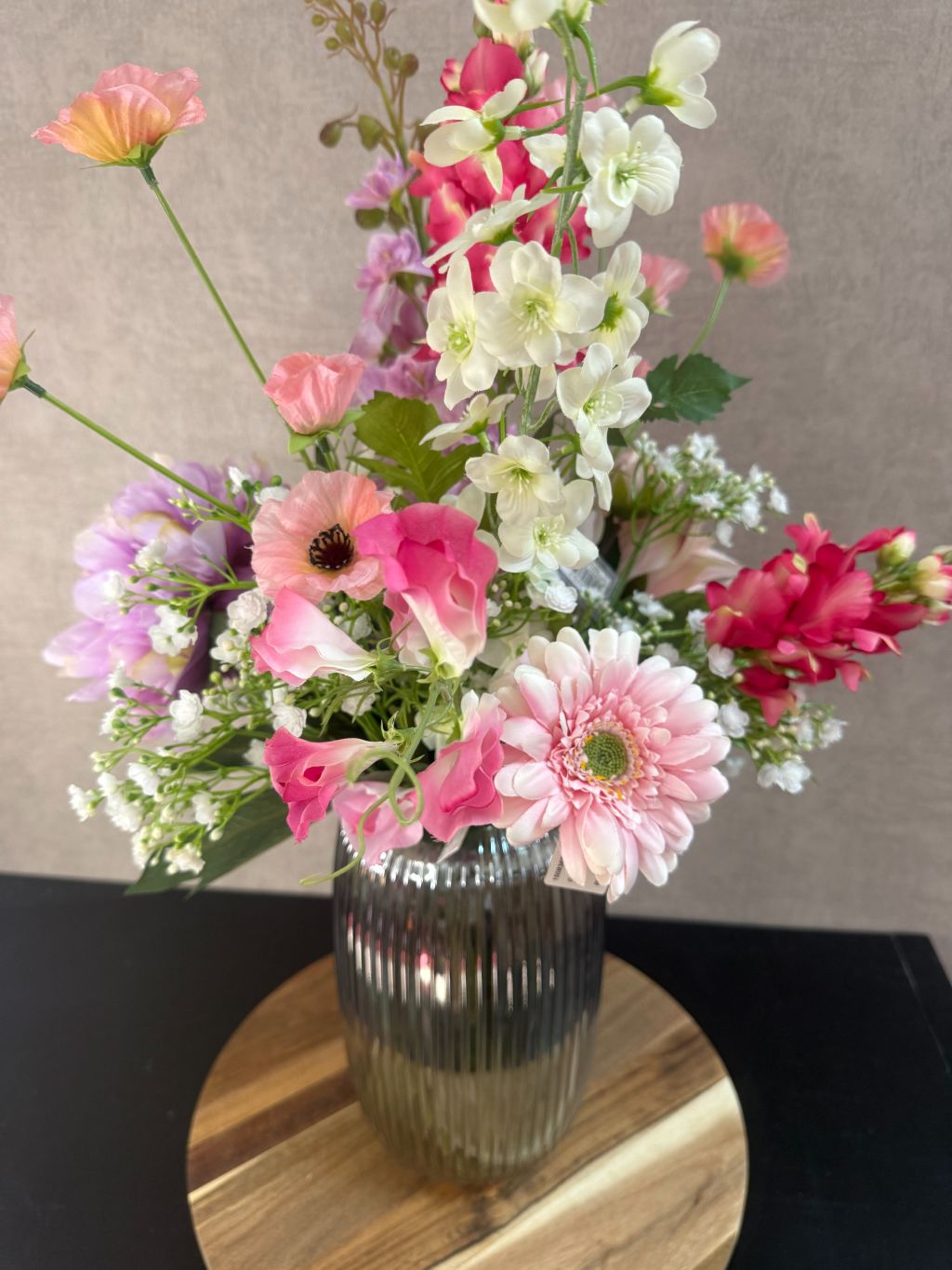Kunstboeket Lovey met paarse, roze en witte bloemen in geribbelde glazen vaas op ronde houten onderzetter.
