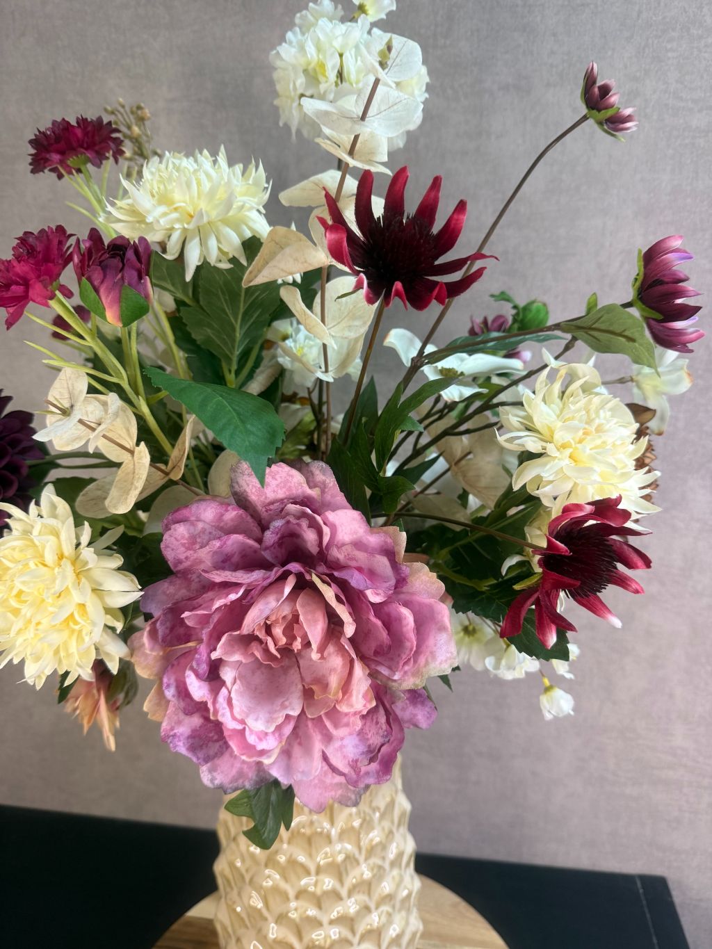 Kunstboeket Noëlle – volle bos kunstbloemen in beige, bruin en bordeaux tinten in een beige vaas tegen grijze achtergrond.