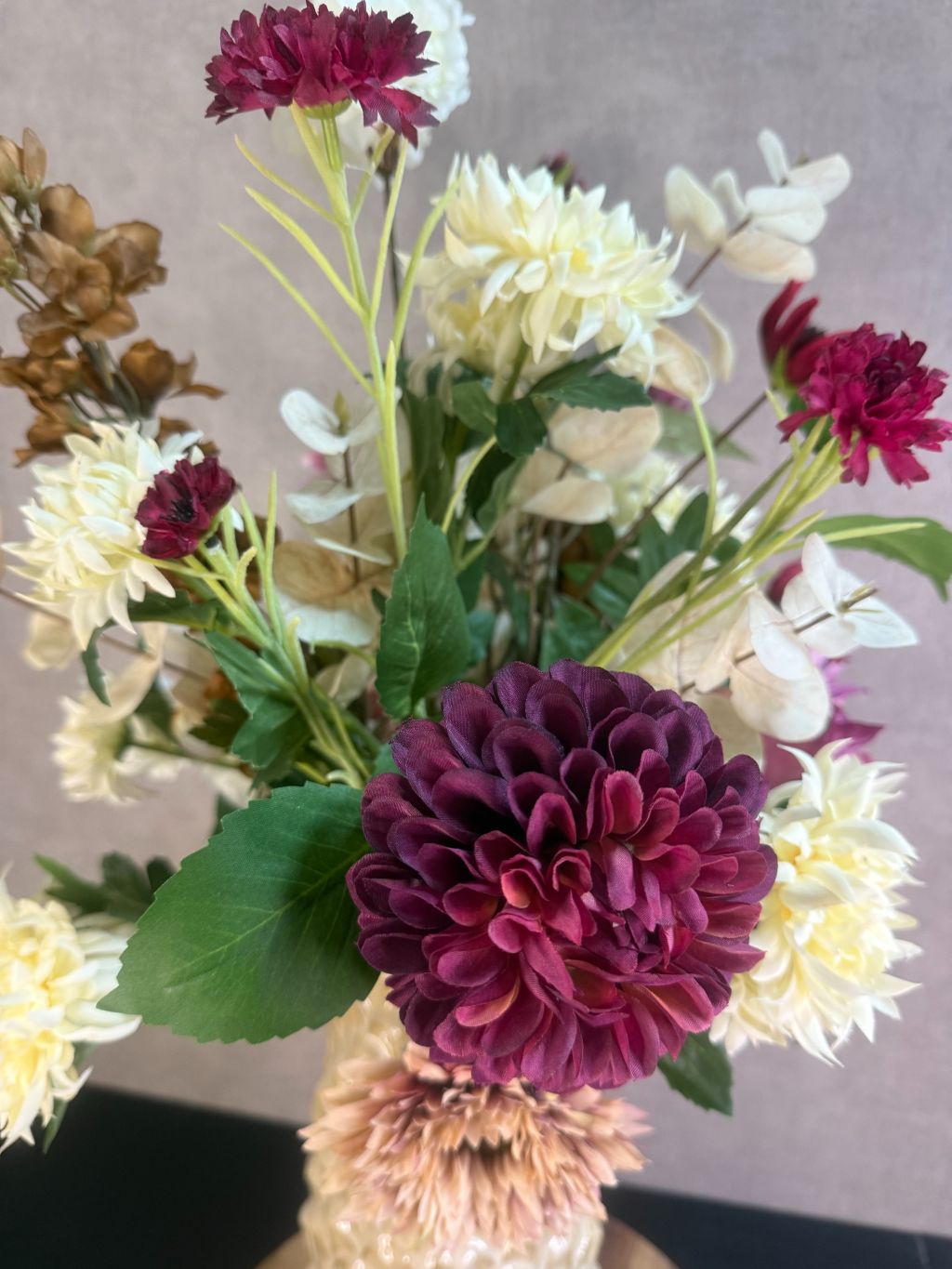 Kunstboeket Noëlle – volle bos kunstbloemen in beige, bruin en bordeaux tinten in een beige vaas tegen grijze achtergrond.