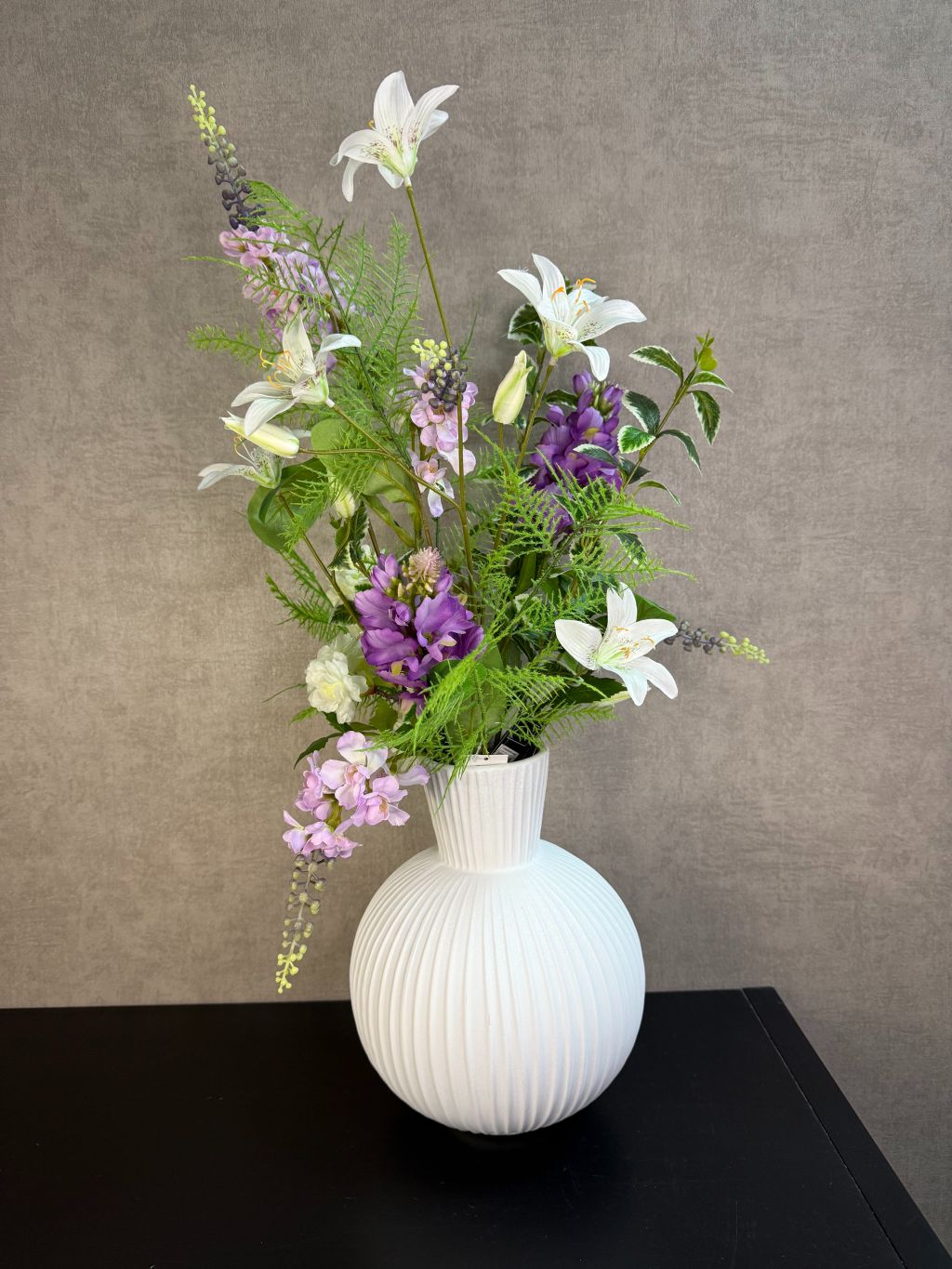 Paars en crèmekleurig kunstboeket met groene takken en witte bloemen in een witte ribbelvaas op een donkere tafel.