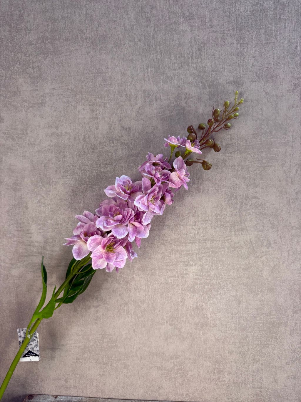 Kunstbloem Delphinium in lila/roze tinten met een volle bloeiwijze en groene steel, liggend op een grijze achtergrond.
