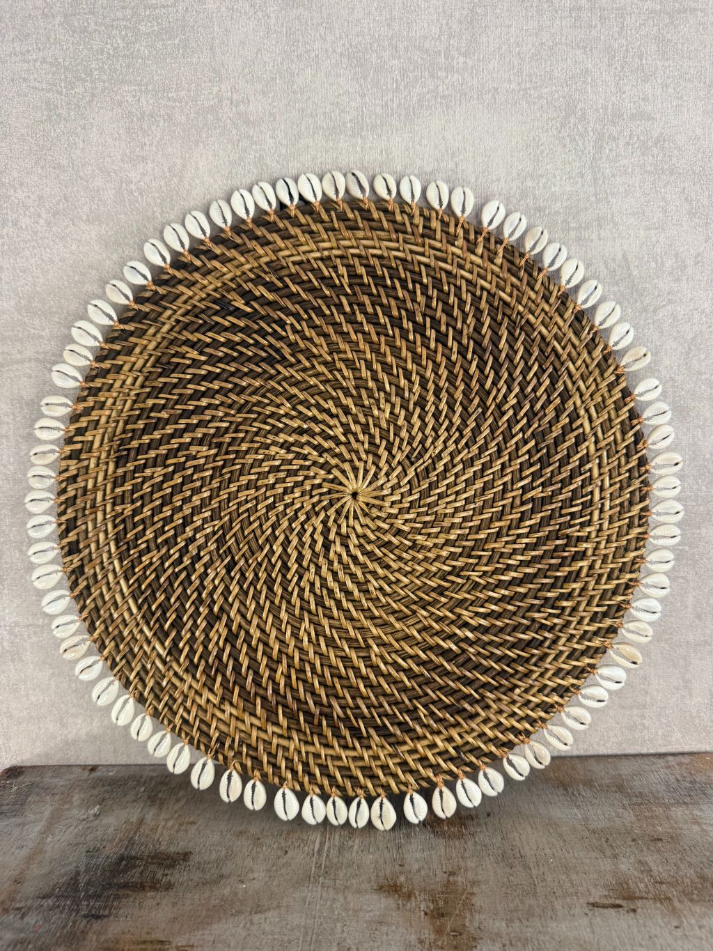 Ronde rotan placemat met witte schelprand, gebruikt als decoratief onderbord of dienblad op houten ondergrond.
