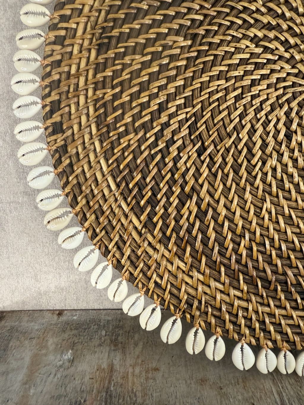 Ronde rotan placemat met witte schelprand, gebruikt als decoratief onderbord of dienblad op houten ondergrond.
