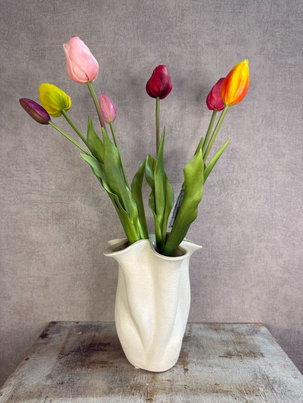 Bos real touch tulpen in een bonte mix van kleuren met groene bladeren, staand op een houten ondergrond tegen een neutrale achtergrond. met vaas
