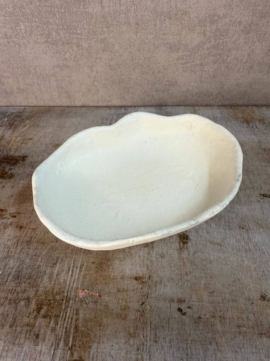 Crèmekleurig keramieken schaaltje met organische, golvende rand, geplaatst op een houten ondergrond tegen een neutrale achtergrond.