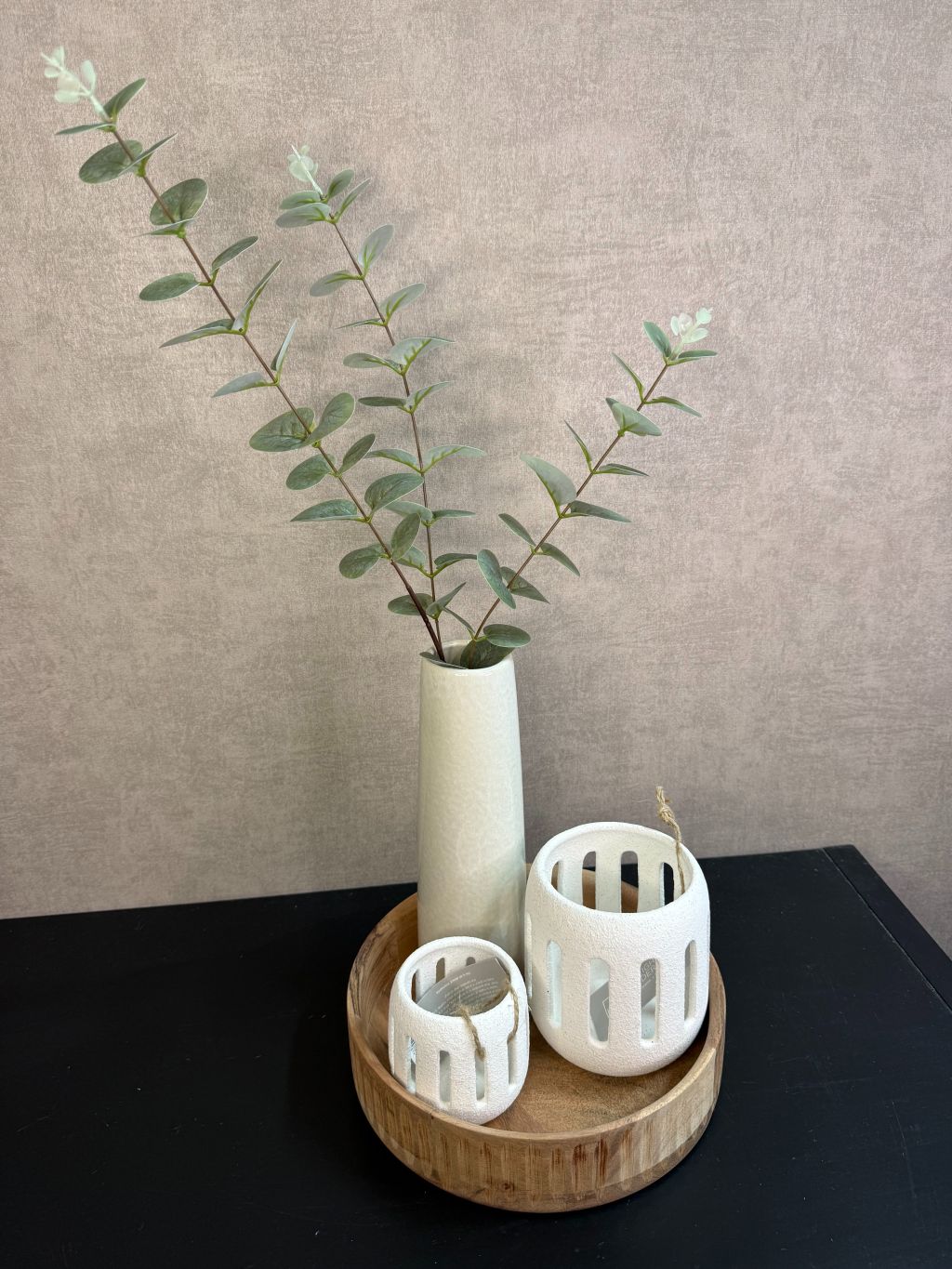 Rond houten dienblad met crème vaas met groene takken en twee crème waxinelichthouders op een zwarte tafel.