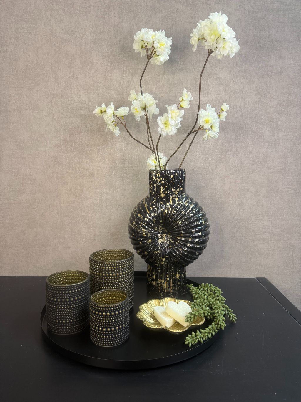 Tafel decoratie set Nelle – zwarte vaas met witte bloemen, goudkleurige schaal met amberblokjes en zwarte waxinelichthouders op rond dienblad.