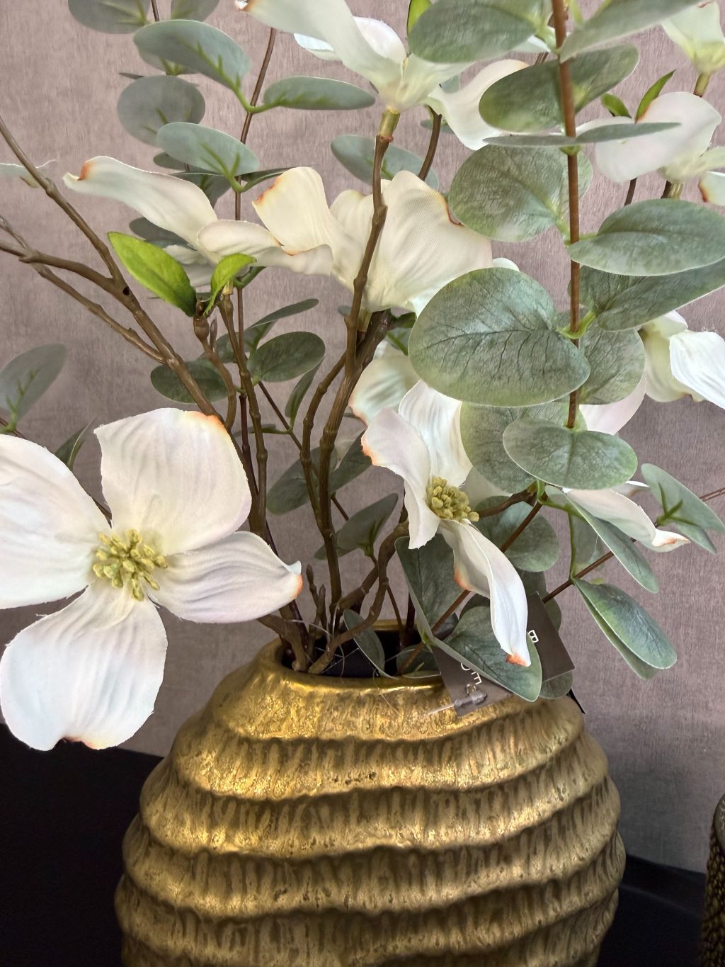 Zwart metalen dienblad met goudkleurige vaas met witte kunstbloemen en twee goud-zwarte waxinelichthouders op een zwarte tafel.