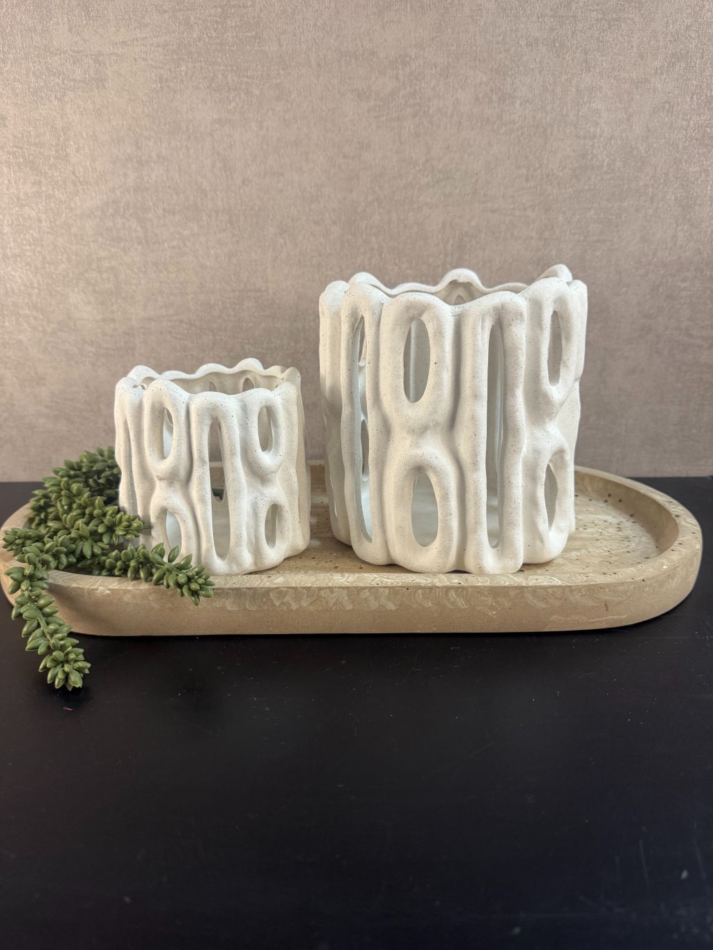 Tafeldecoratieset met zandkleurig dienblad, twee witte sculpturale waxinelichthouders en groen hangplantje op zwarte ondergrond.