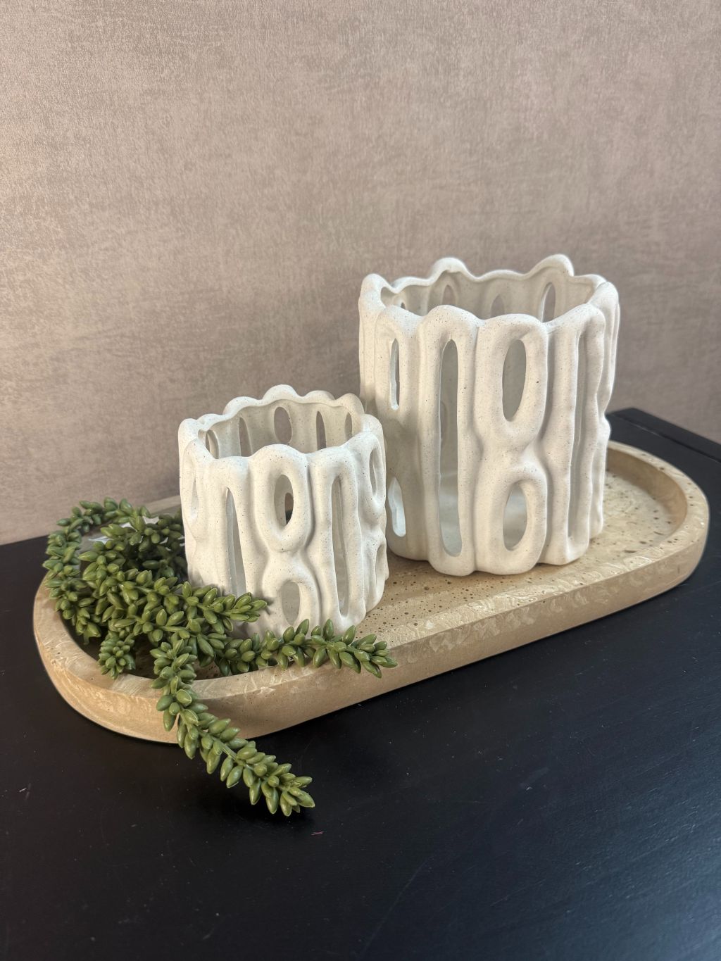 Tafeldecoratieset met zandkleurig dienblad, twee witte sculpturale waxinelichthouders en groen hangplantje op zwarte ondergrond.