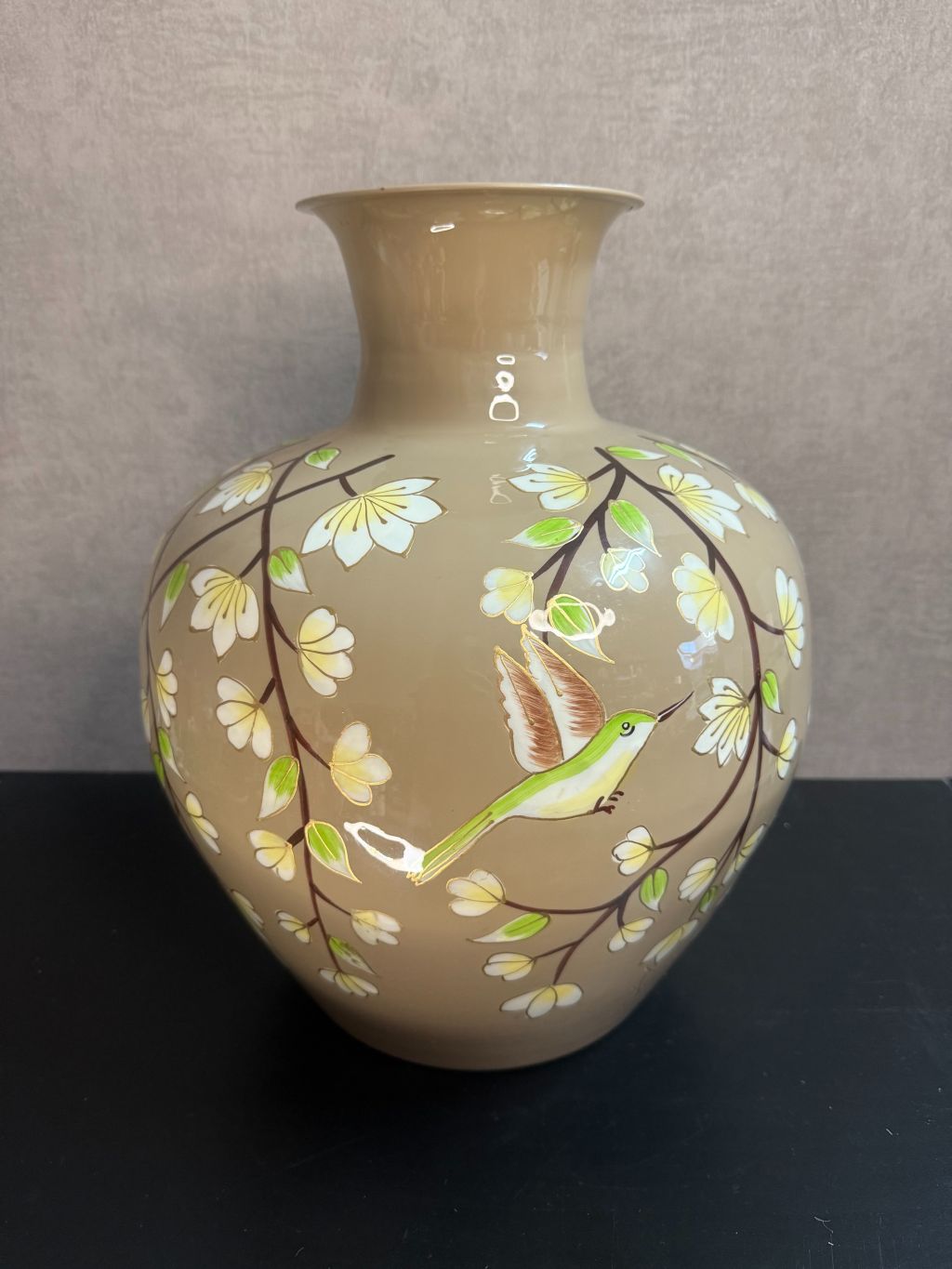 Handgeschilderde metalen vaas in taupe met bloemen en kolibrie in wit, groen en goud op een zwarte ondergrond.