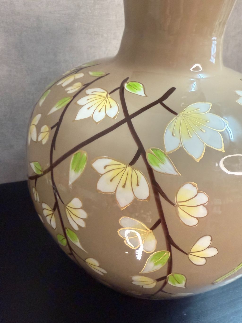 Handgeschilderde metalen vaas in taupe met bloemen en kolibrie in wit, groen en goud op een zwarte ondergrond.