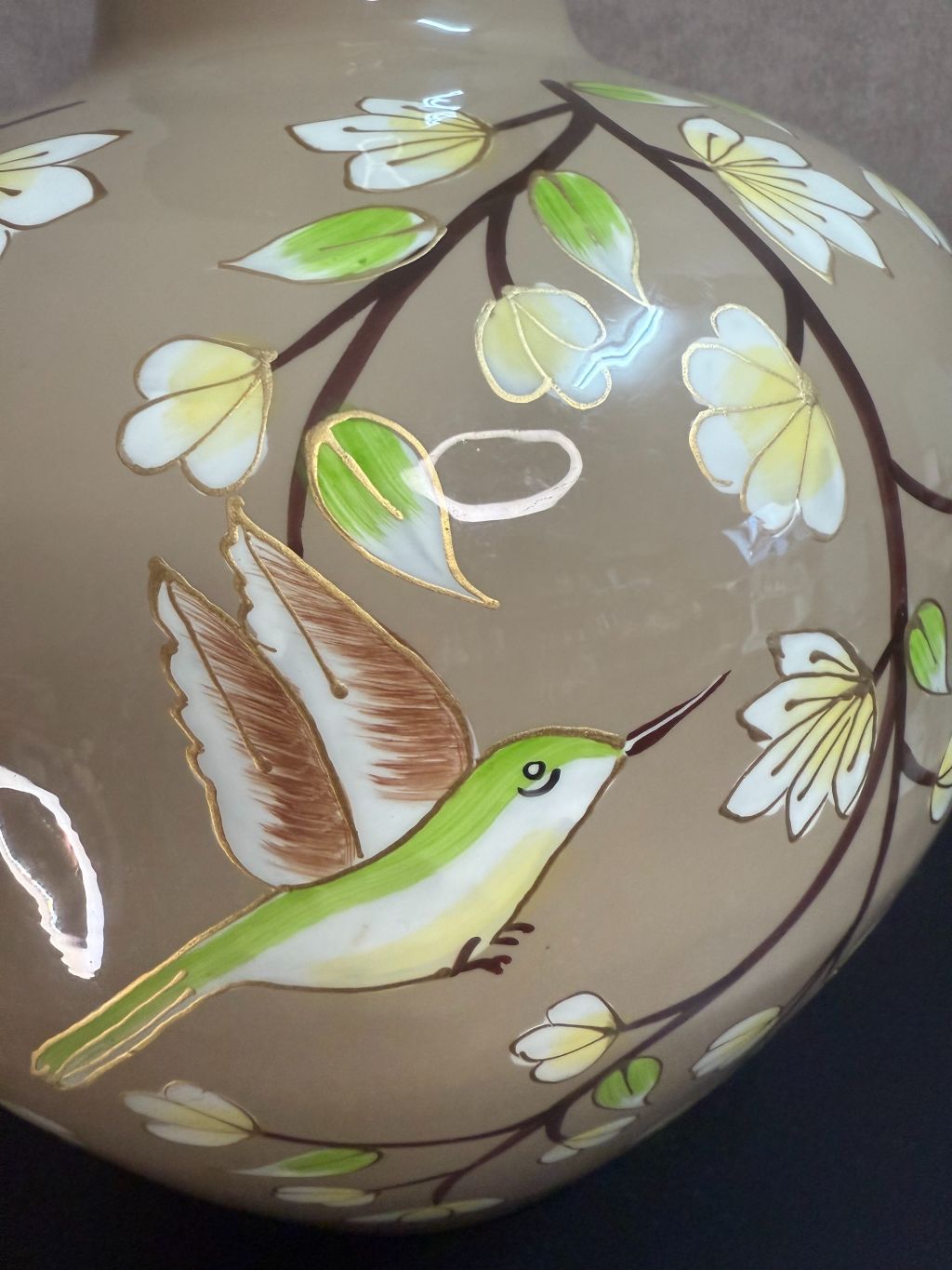 Handgeschilderde metalen vaas in taupe met bloemen en kolibrie in wit, groen en goud op een zwarte ondergrond.