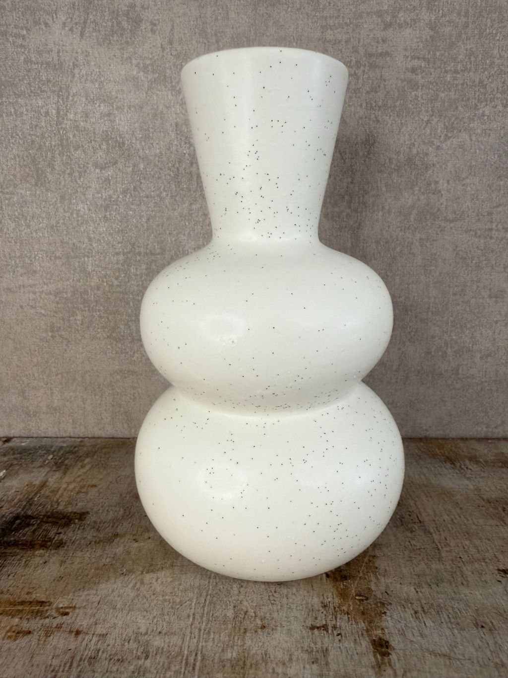 Witte keramieken vaas met zwarte spikkels en bolvormig design, staand op een houten oppervlak tegen een grijze achtergrond.