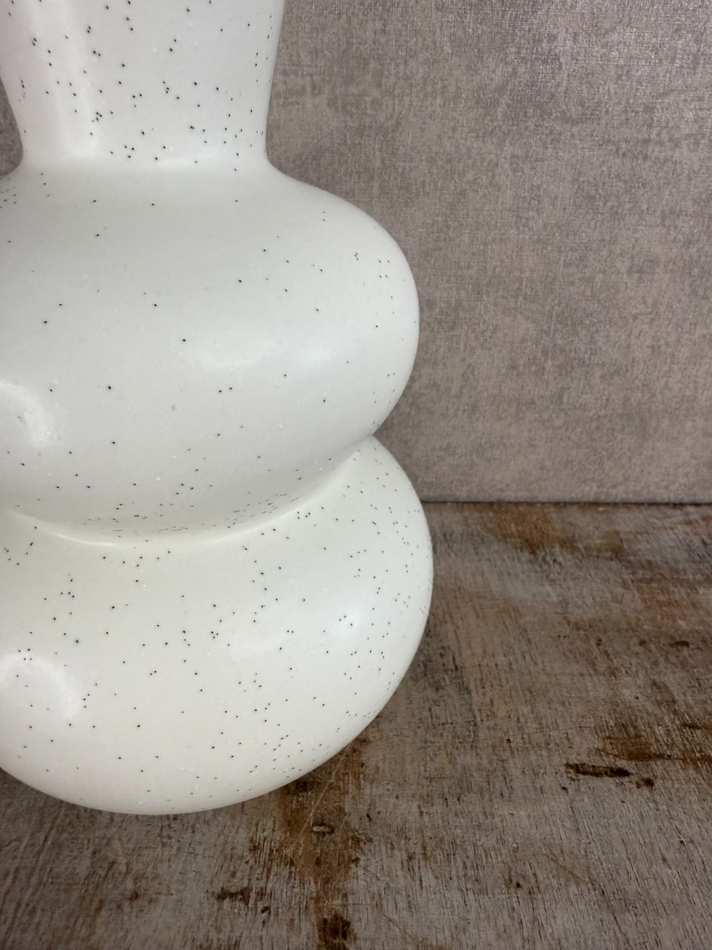 Witte keramieken vaas met zwarte spikkels en bolvormig design, staand op een houten oppervlak tegen een grijze achtergrond.