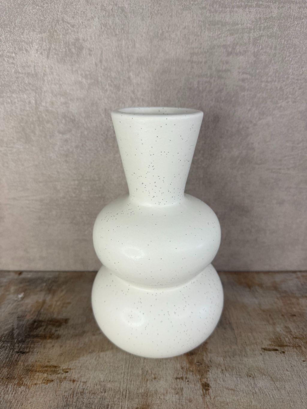 Witte keramieken vaas met zwarte spikkels en bolvormig design, staand op een houten oppervlak tegen een grijze achtergrond.