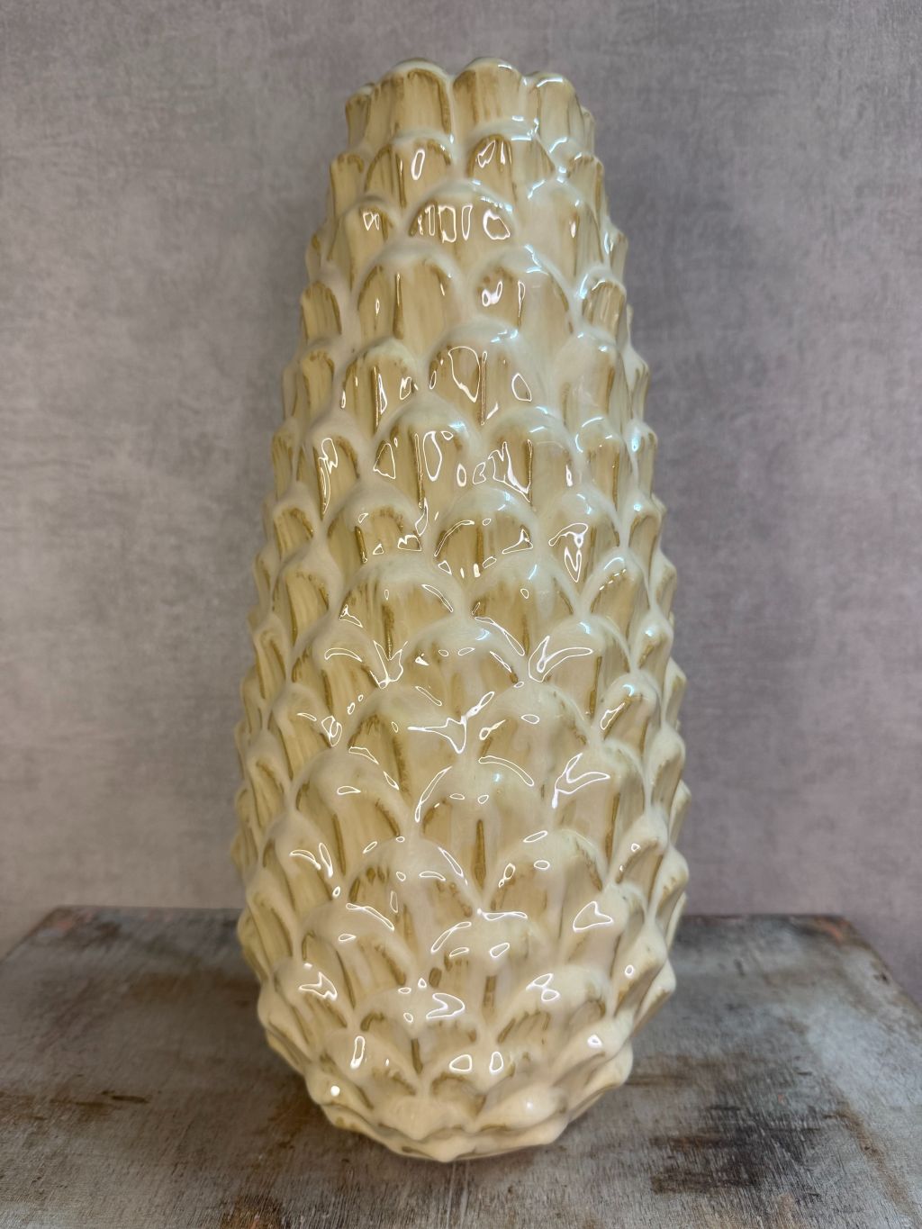 Vaas Pinecone beige – keramieken vaas met glanzende dennenappelstructuur op een houten ondergrond tegen een grijze muur.