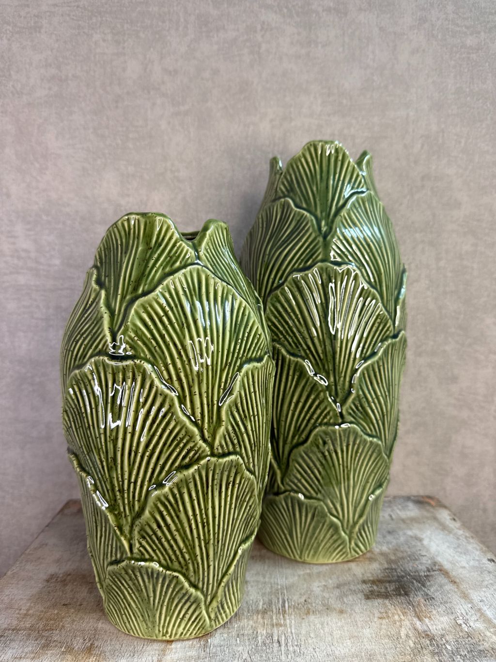 Groene keramieken vazen met bladstructuur in twee formaten op een houten ondergrond tegen een neutrale achtergrond.
