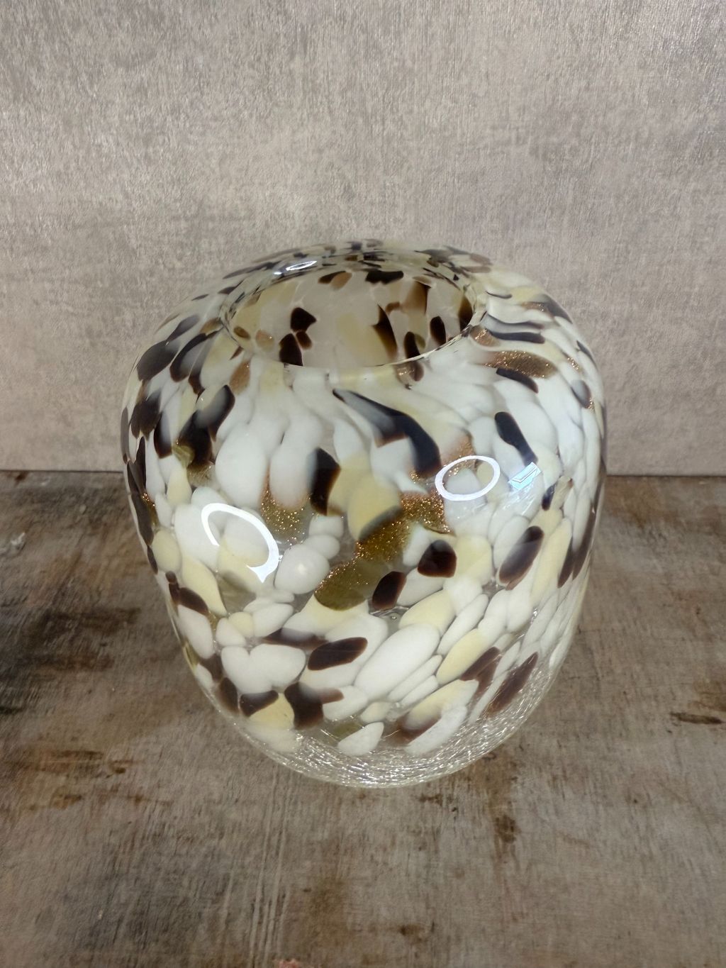 Glazen vaas met gevlekt patroon in beige, wit, bruin en goud, staand op houten oppervlak tegen een neutrale achtergrond.