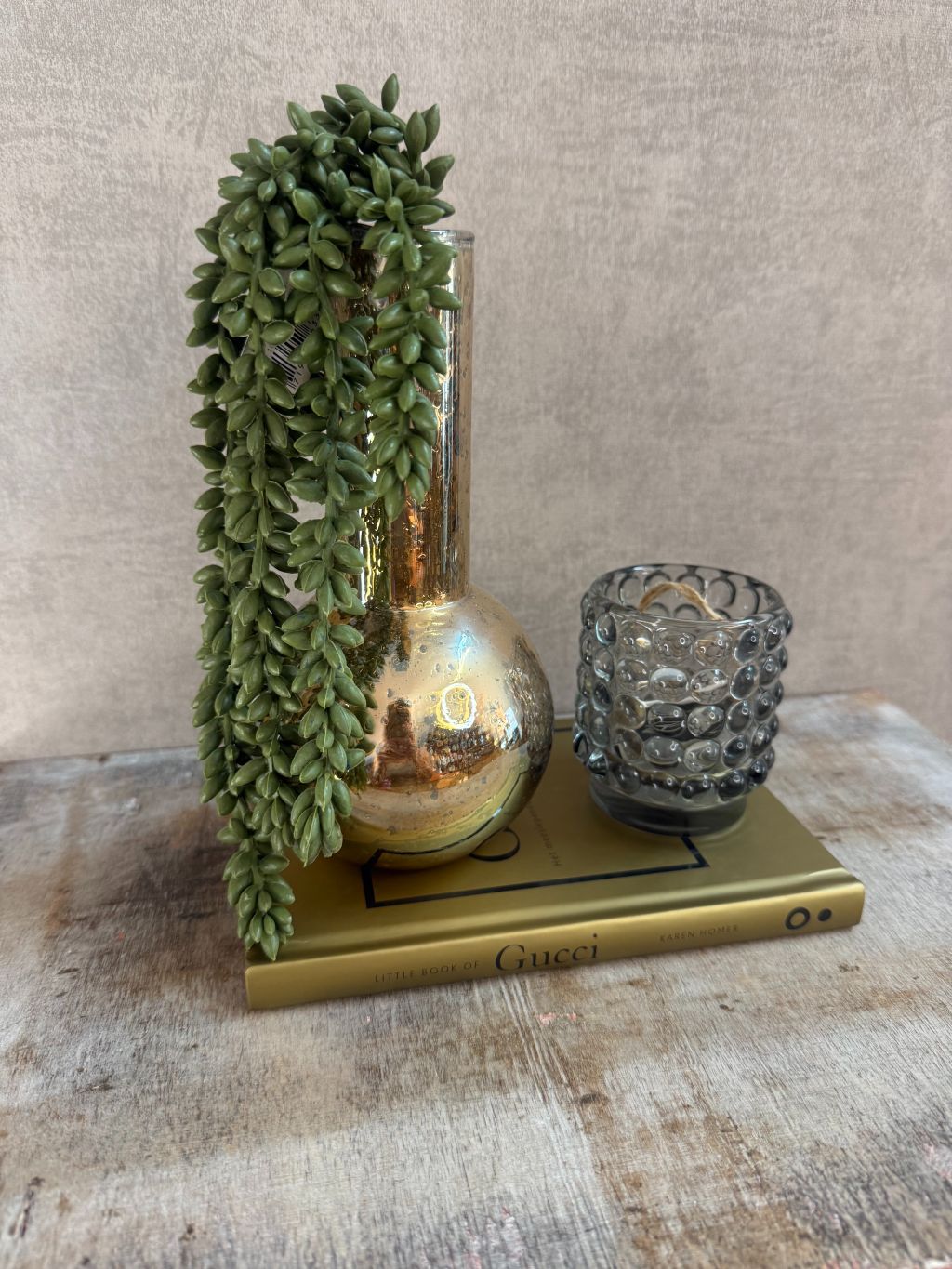 Set met gouden vaas, groene hangplant, smokey waxinelichthouder en Gucci boek op houten ondergrond.