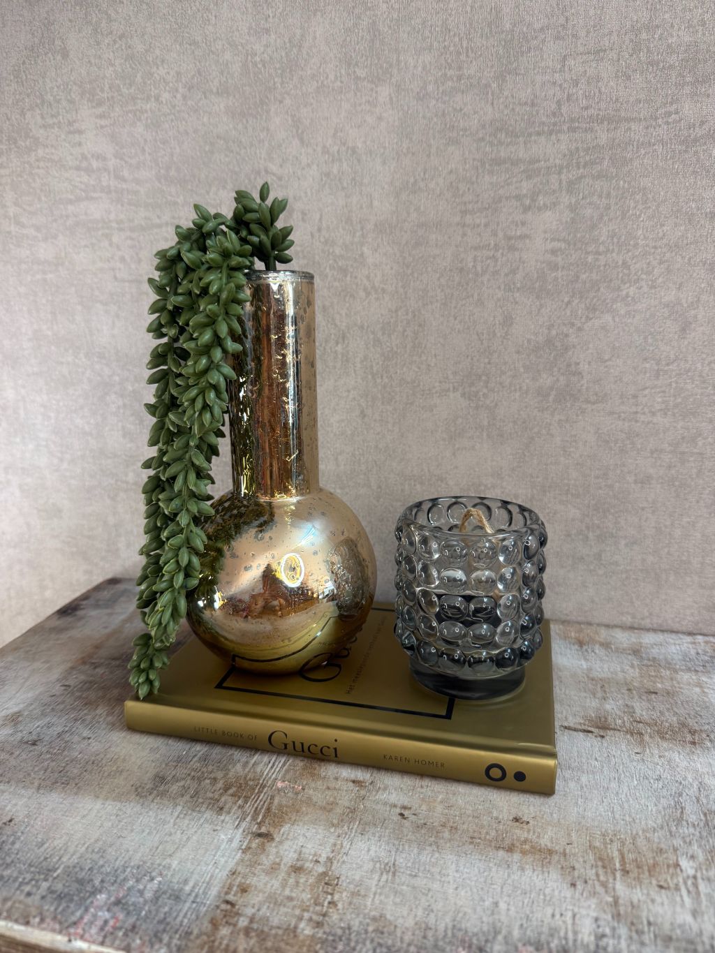 Set met gouden vaas, groene hangplant, smokey waxinelichthouder en Gucci boek op houten ondergrond.