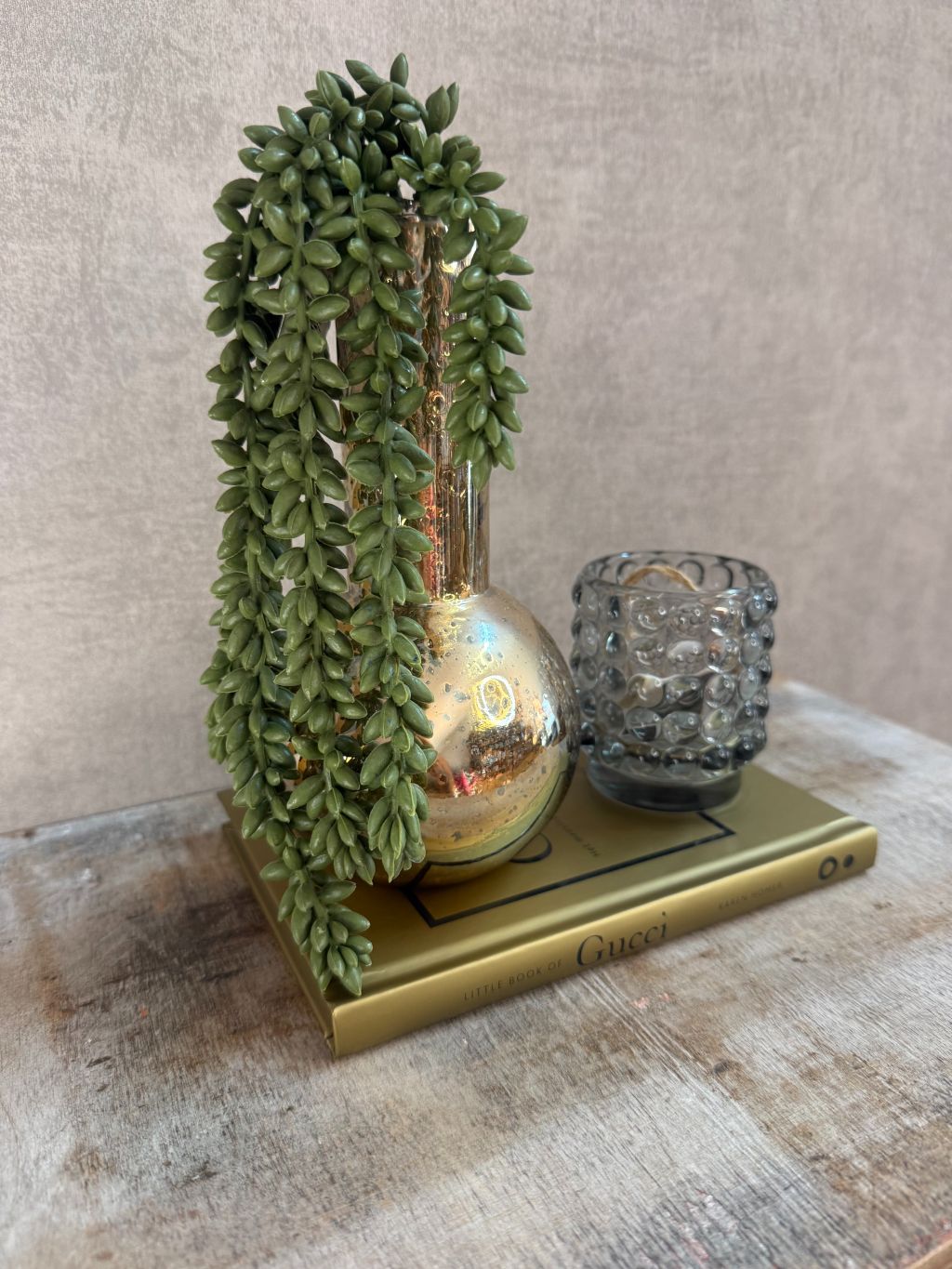 Set met gouden vaas, groene hangplant, smokey waxinelichthouder en Gucci boek op houten ondergrond.