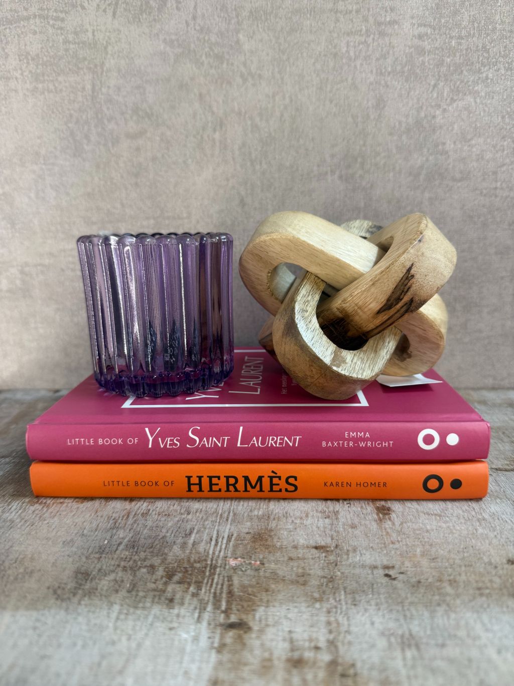 Decoratieset met Hermès- en YSL-boek, paarse glazen waxinelichthouder en houten knoopornament op houten ondergrond.