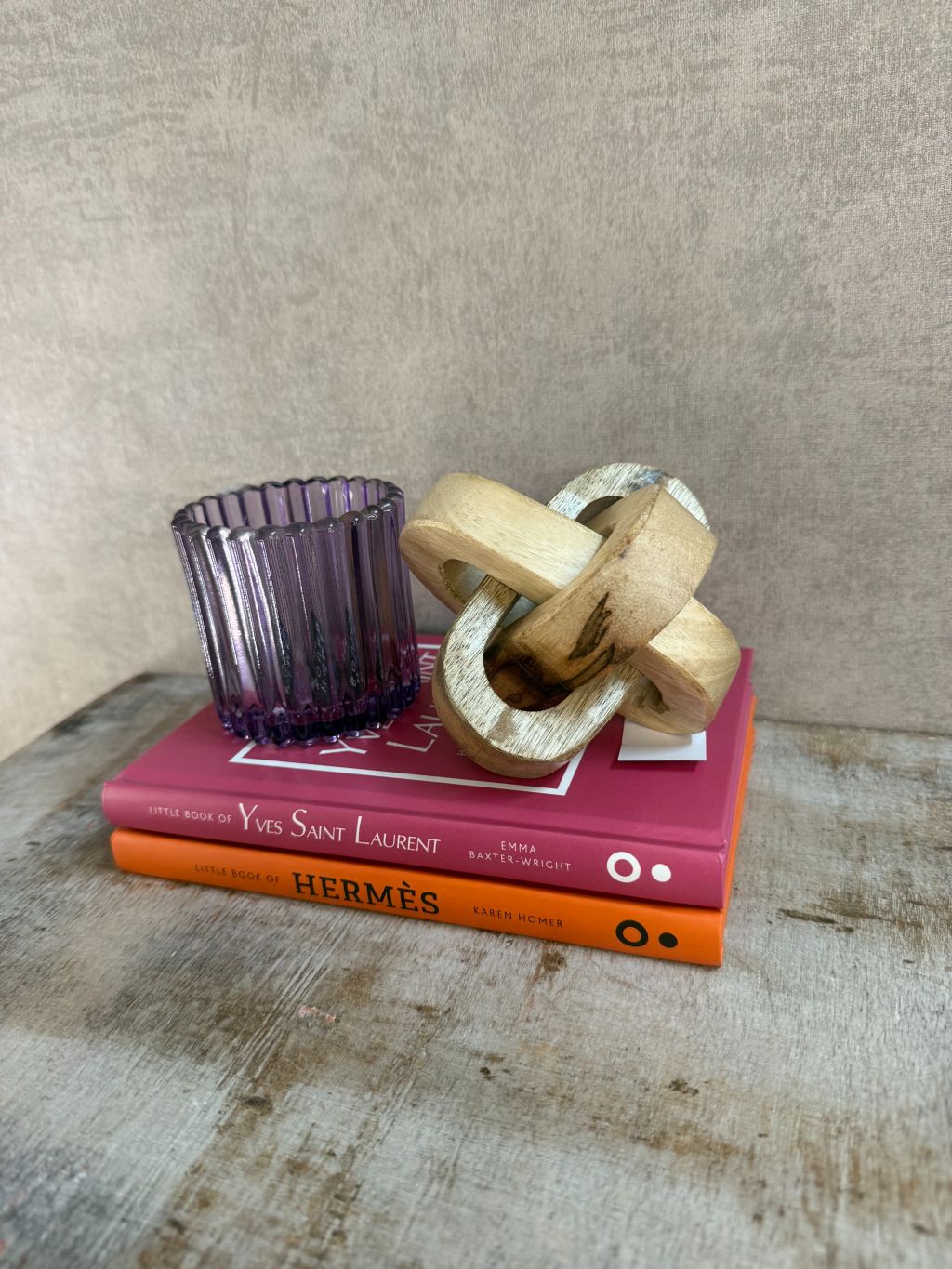 Decoratieset met Hermès- en YSL-boek, paarse glazen waxinelichthouder en houten knoopornament op houten ondergrond.