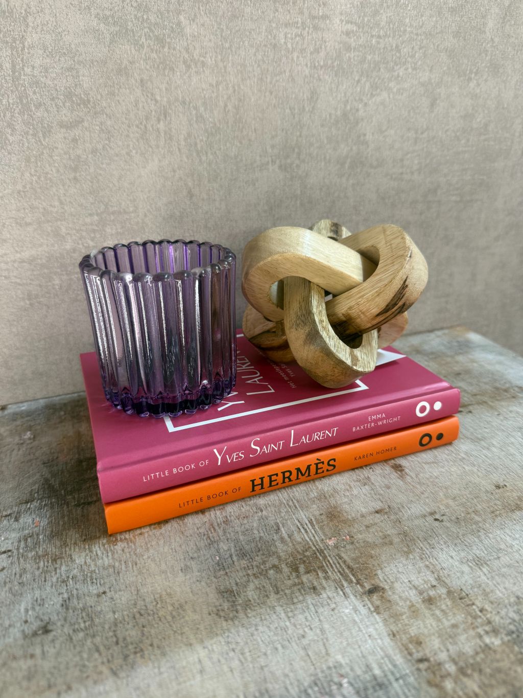 Decoratieset met Hermès- en YSL-boek, paarse glazen waxinelichthouder en houten knoopornament op houten ondergrond.